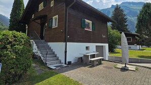 Une maison en bois à deux étages, dotée d’un escalier, d’un petit espace extérieur aménagé pour s’asseoir et offrant une vue sur les montagnes.