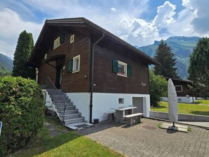 Une maison en bois à deux étages, dotée d’un escalier, d’un petit espace extérieur aménagé pour s’asseoir et offrant une vue sur les montagnes.