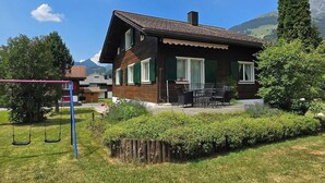 Une maison à deux étages avec un patio, entourée de verdure et avec des montagnes en arrière-plan.