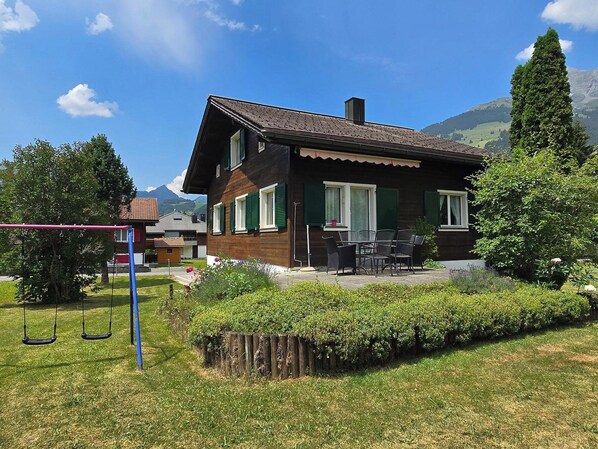 Une maison à deux étages avec un patio, entourée de verdure et avec des montagnes en arrière-plan.