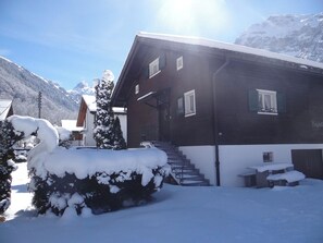 A two-story wooden house with a staircase leading to the entrance, surrounded by snow-covered trees and mountains in the background. - Chalet Fluegärtli Appartement/Fewo 6-Bettwohnung by Interhome (Engelberg)