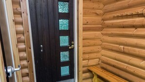 Puisesta hirsirakenteesta tehty mökki, jossa on tumma ovi, jossa on lasiruudut, kivilaatoitettu sisäänkäynti ja puinen penkki.