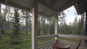 Une terrasse en bois avec une table et des chaises, donnant sur une zone boisée avec de grands arbres et un champ d’herbe.