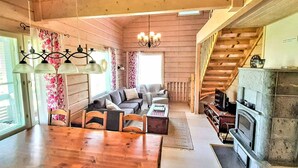 L’intérieur chaleureux d’un chalet avec une table à manger en bois, un ensemble de canapés, une cheminée et un escalier.