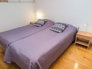 Una cama doble con colcha púrpura acolchada y almohadas a juego, una mesita de noche de madera con lámpara, y un radiador en la pared.