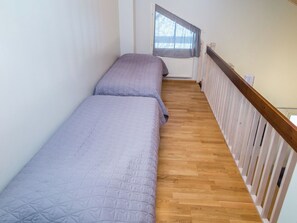 Um corredor estreito com duas camas, soalho de madeira e uma janela com cortinas.