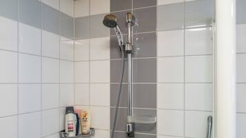 A shower with a handheld showerhead and a wall-mounted faucet. There is a shelf with toiletries.