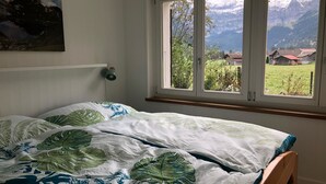A bedroom with a bed, a window with a view of mountains and a village, and a wall-mounted lamp.