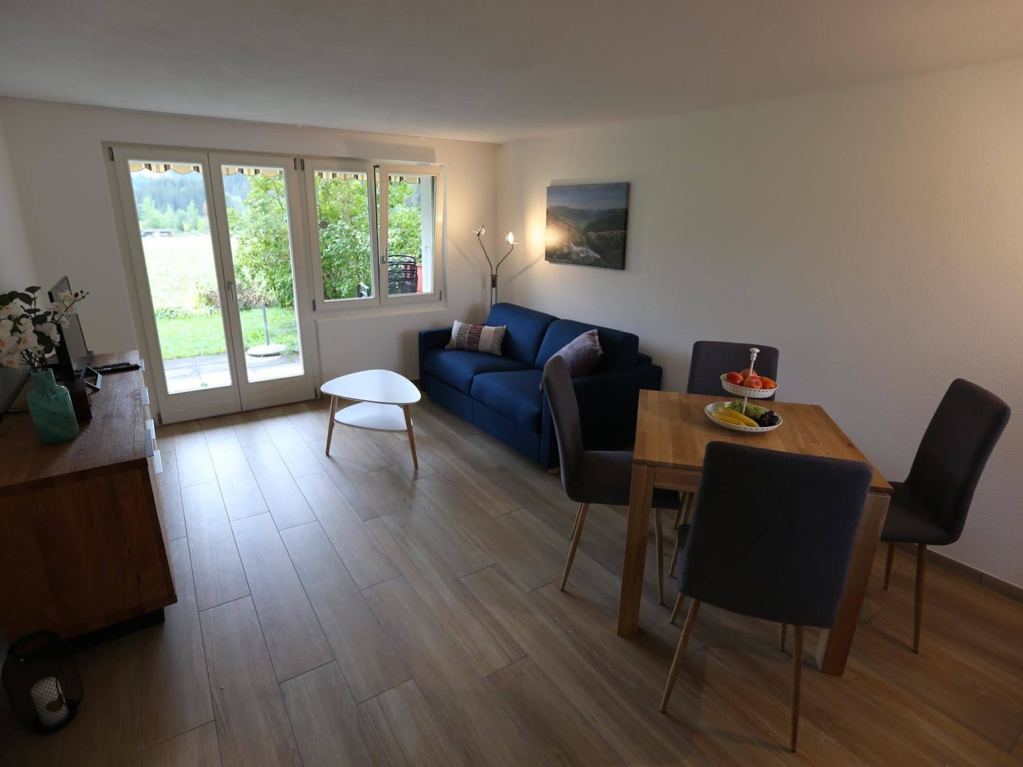 A modern living room with a sofa, a dining table with chairs, a small white table, and a large window with a view of greenery.