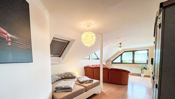 Un dormitorio tipo loft moderno con techo inclinado, sofá empotrado, ventilador de techo y ventanas amplias.