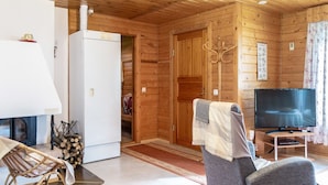 Interno accogliente di una baita con arredamento in legno, un frigorifero bianco, una televisione su un mobile, una sedia e un tavolino.