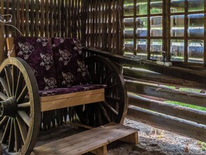 A rustic wooden bench with a floral cushion, a large wooden wheel, and a wooden table in a room with a wooden fence and windows. - Pikkupuro by Interhome (Nilsiä)