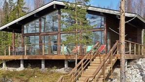 Puinen mökki, jossa on suuret ikkunat, puinen terassi ja portaat, jotka johtavat sisäänkäynnille.