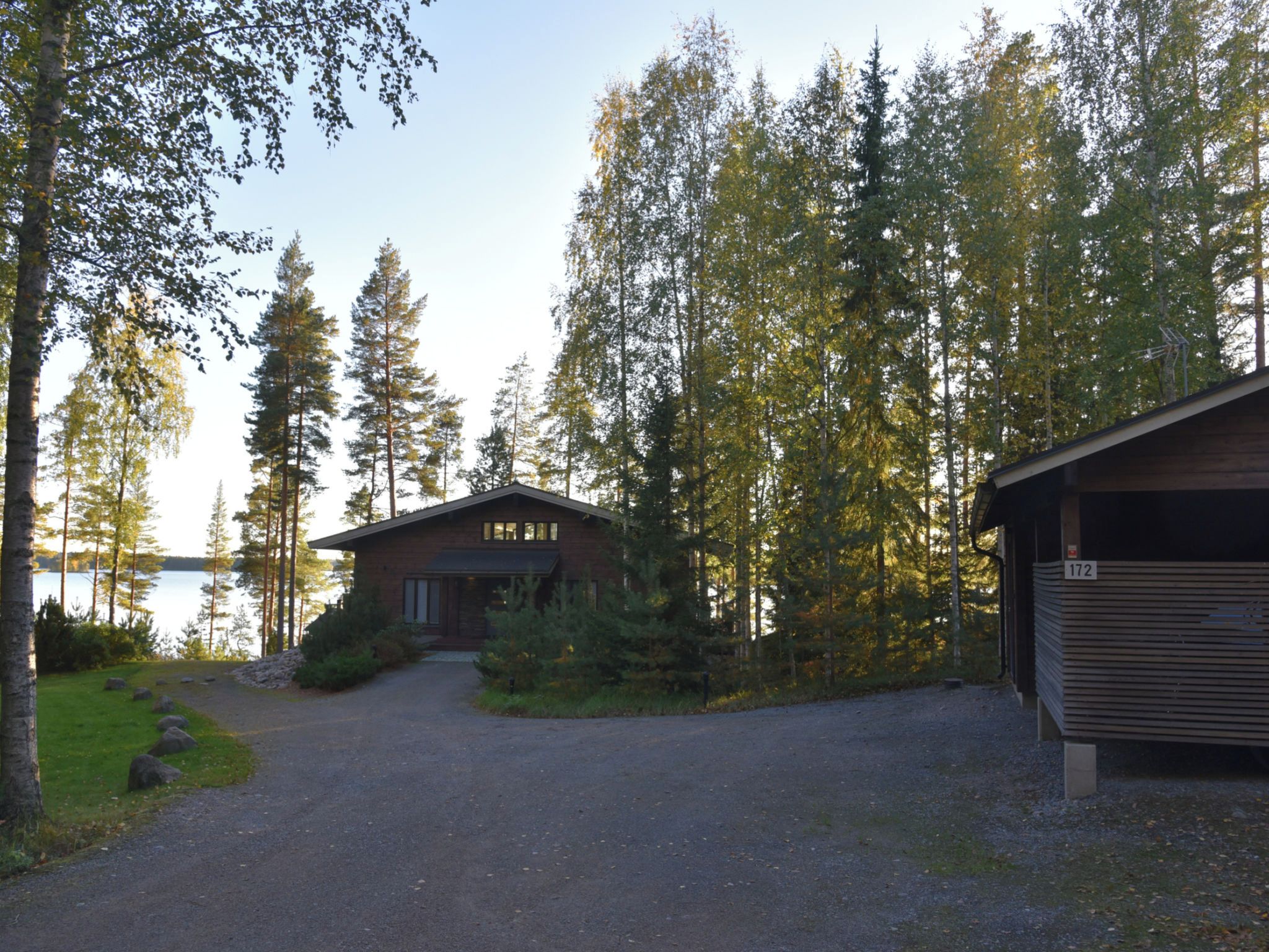 Une cabane avec une allée de gravier menant à une porte en bois, entourée d’une forêt dense et avec une étendue d’eau en arrière-plan.