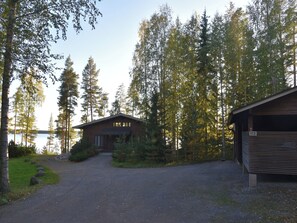 A cabin with a gravel driveway leading to a wooden door, surrounded by dense forest and a body of water in the background.