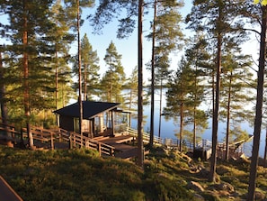En træhytte med terrasse, omgivet af høje fyrretræer og med udsigt over vandet.