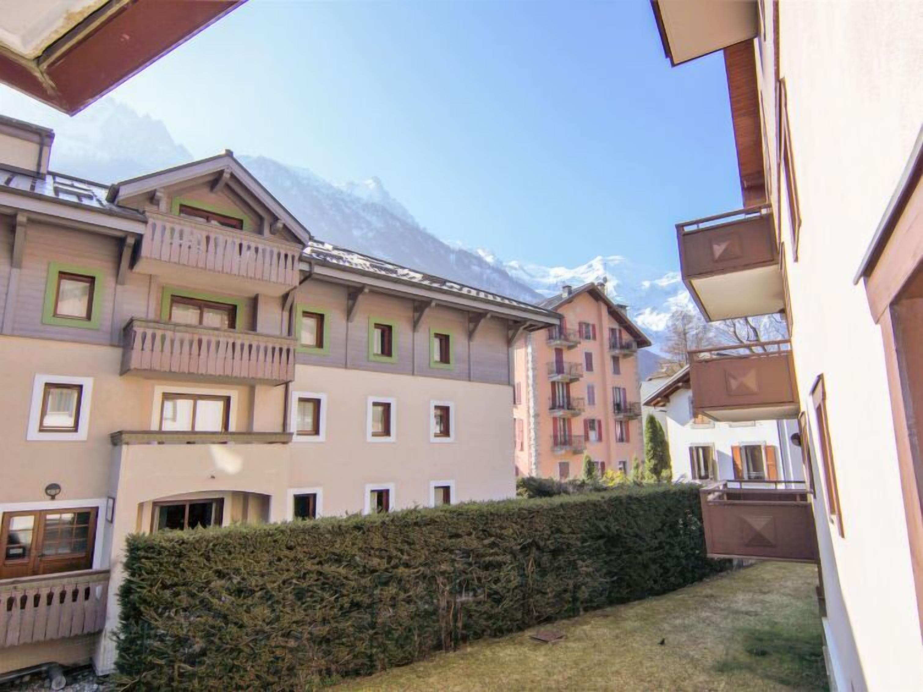 A multi-story building with balconies and windows, surrounded by a hedge and a mountain in the background.