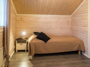 A small, cozy room with a wooden slat ceiling, a single bed with a brown bedspread, a bedside table with a lamp, and a window with curtains.