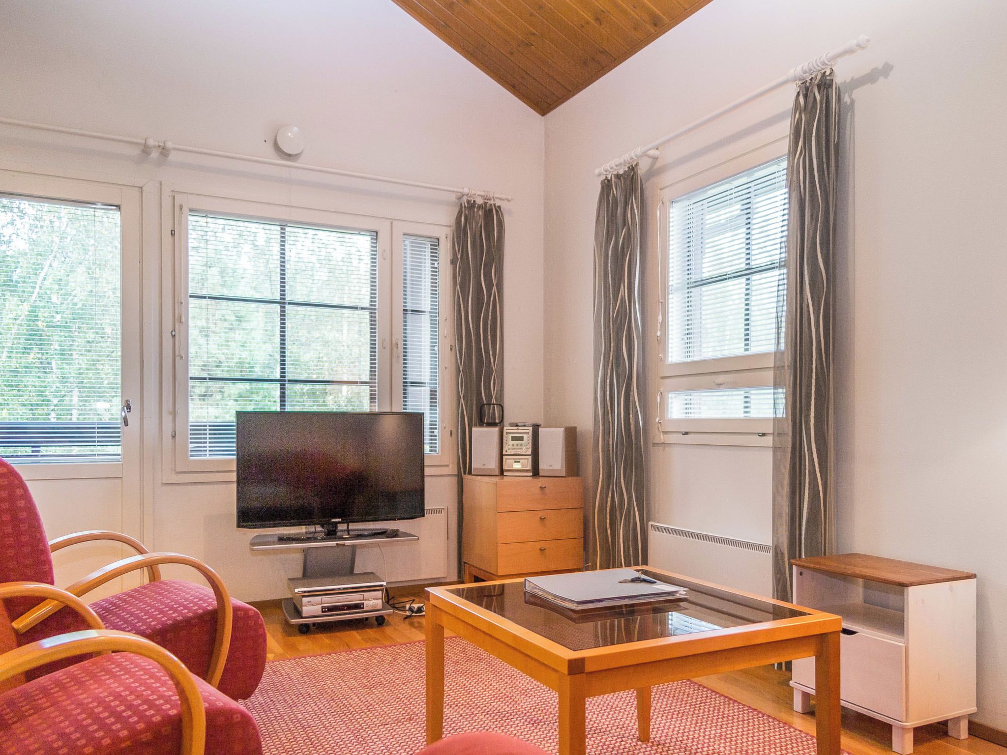 Ein Zimmer mit Fernseher, einem Holztisch, einem roten Sessel und einem Fenster mit Vorhängen.