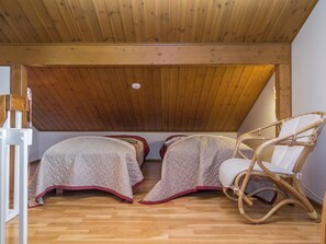 Una habitación pequeña y acogedora con techo inclinado, dos camas con colchas, una silla de mimbre y un radiador blanco.