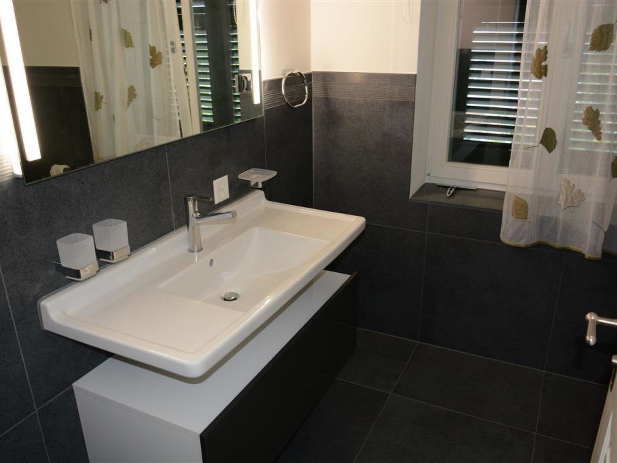 A modern bathroom with a white sink, dark tiles, a mirror, and a window with leaf-patterned curtains.
