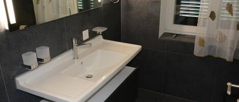 A modern bathroom with a white sink, dark tiles, a mirror, and a window with leaf-patterned curtains.