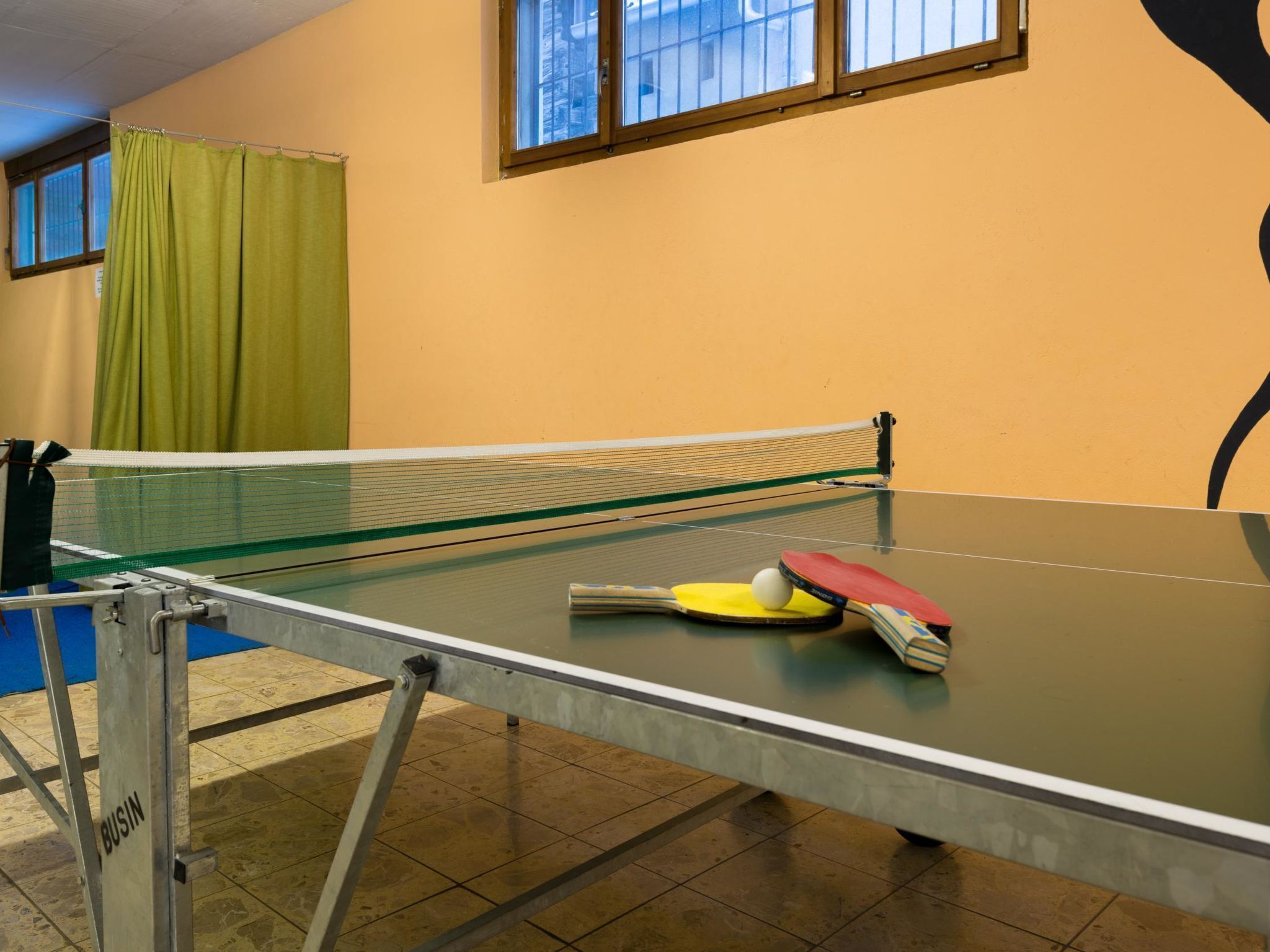Une table de ping-pong avec des raquettes et une balle, dans une pièce dotée d’un rideau vert et d’une fenêtre.