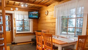 A cozy cabin interior with a wooden dining table, chairs, a television mounted on the wall, and large windows offering a view of the surrounding forest. - Vacation home Varsta by Interhome (Kuusamo)