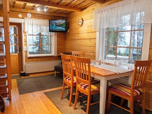 A cozy cabin interior with a wooden dining table, four chairs with colorful cushions, a television mounted on the wall, and large windows with curtains.