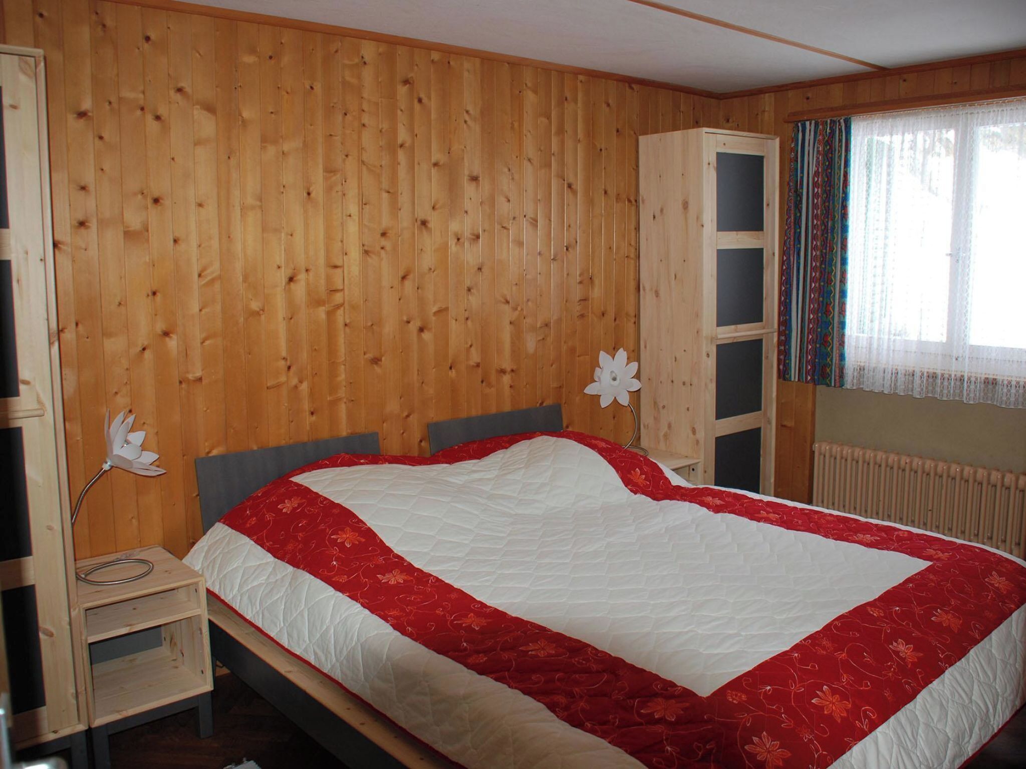 A bedroom with a wooden paneled wall, a bed with a white and red quilt, a nightstand, and a wardrobe.