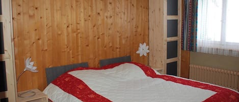 A bedroom with a wooden paneled wall, a bed with a white and red quilt, a nightstand, and a wardrobe.