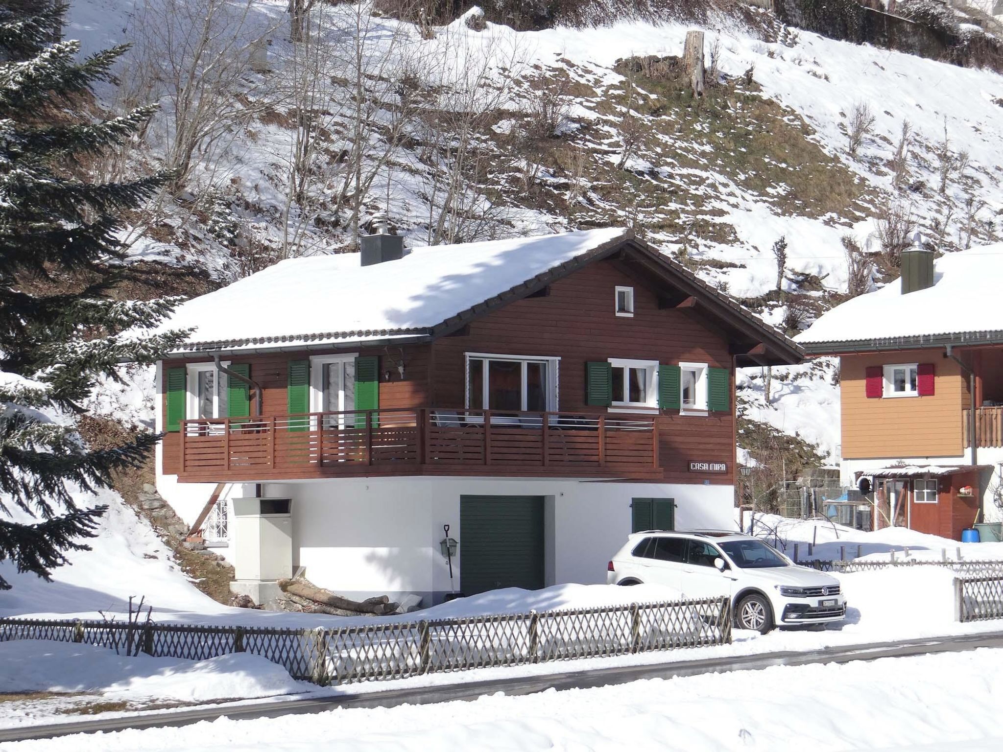 A two-story chalet with a balcony, a green door, and a car parked in front.