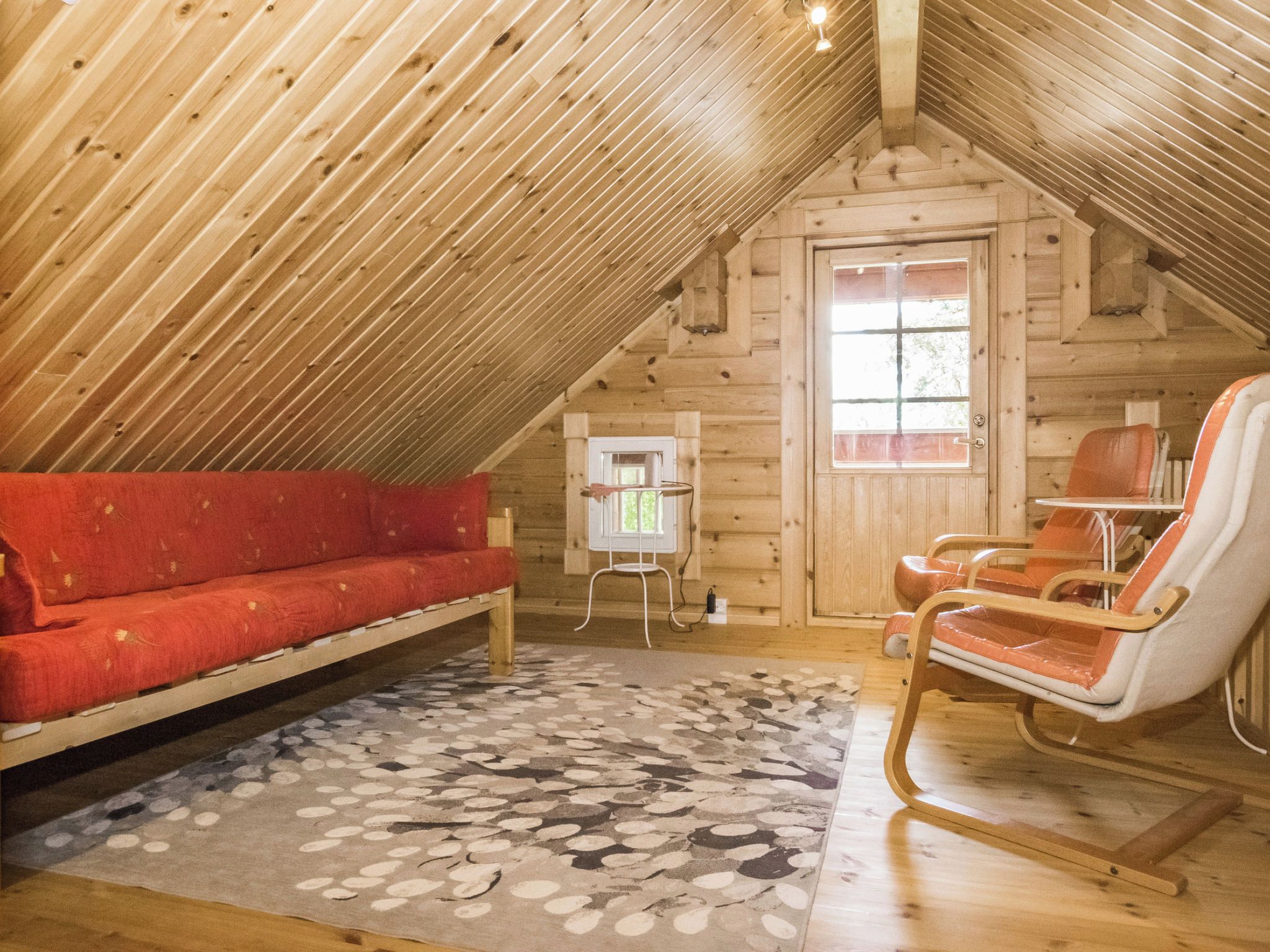 Una habitación cálida en el ático con techo inclinado de madera, un sofá rojo, un sillón mecedor y una alfombra estampada.
