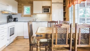 Une cuisine compacte avec des meubles blancs, un four à micro-ondes, un réfrigérateur et une machine à café. On y trouve une table à manger en bois avec des chaises, ainsi qu’une fenêtre avec des rideaux.