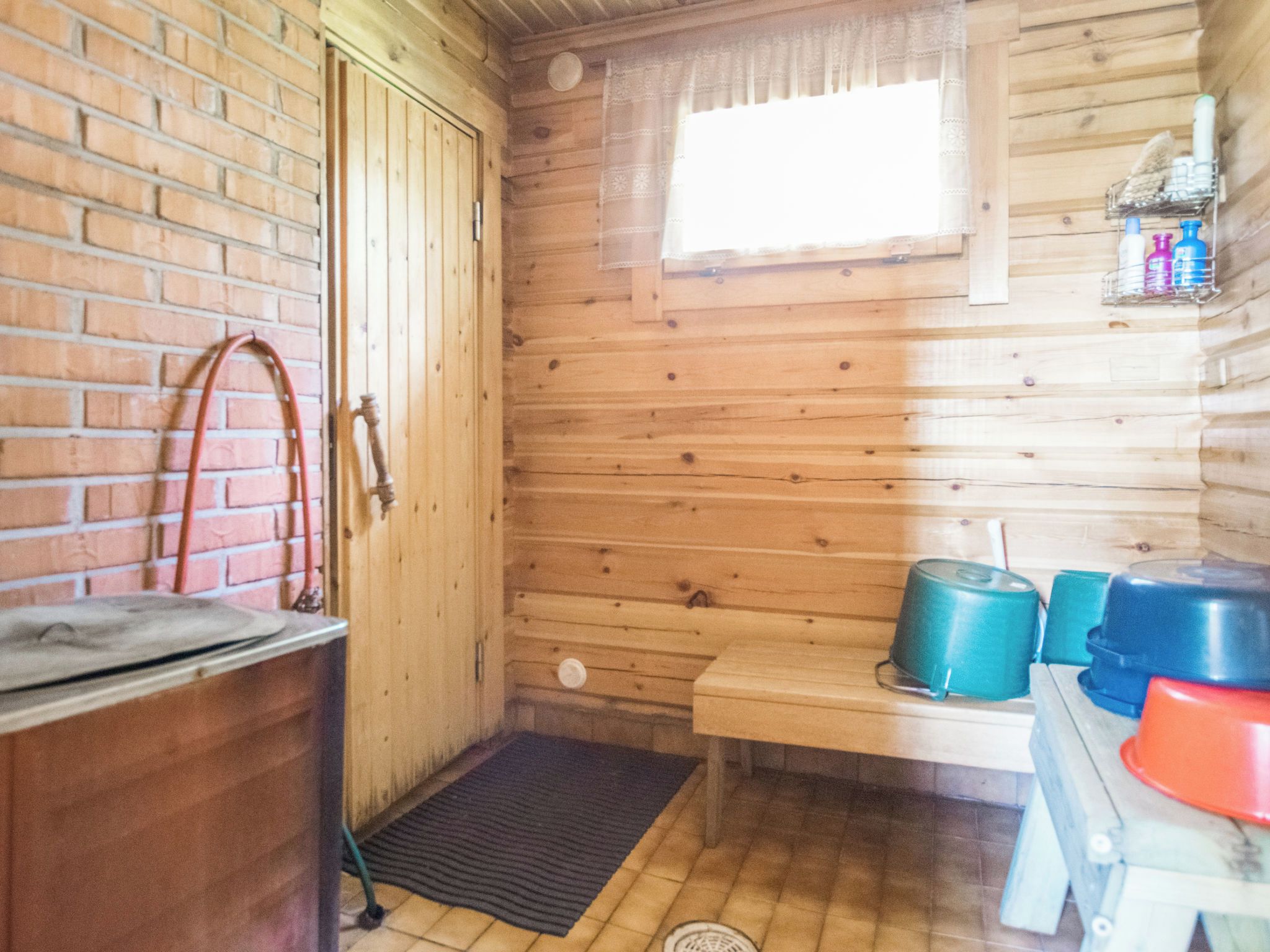 Interior de una cabaña de madera con pared de ladrillo, una puerta de madera, una ventana con cortina, un estante pequeño con botellas, y una mesa con una olla azul y una roja.