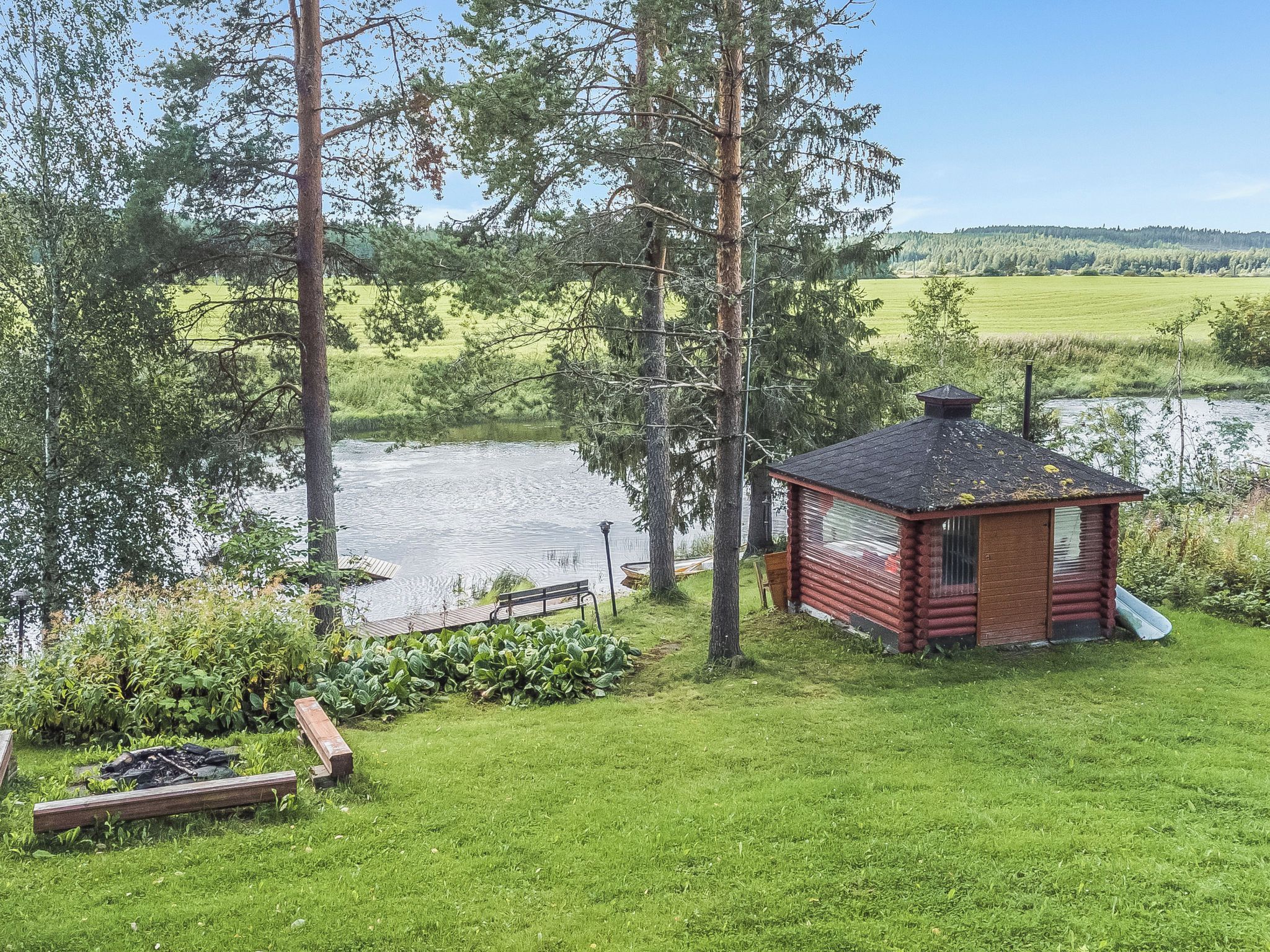 Une cabane en rondins avec une petite terrasse, entourée d’une zone herbeuse, d’arbres et d’une étendue d’eau.