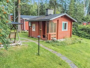 A small red cabin with a porch, surrounded by greenery and a paved path.