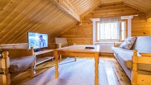 Acogedora habitación en ático con paredes de madera y techo inclinado. Incluye un televisor, un sofá, una mesa y una ventana con cortinas.