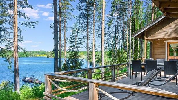 Terraza de madera con mobiliario exterior con vista a un lago, rodeada de árboles altos.