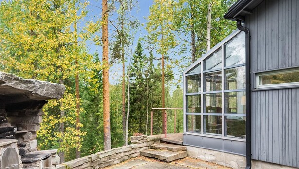 A stone patio with a stone fireplace, surrounded by trees and a modern building with large windows.