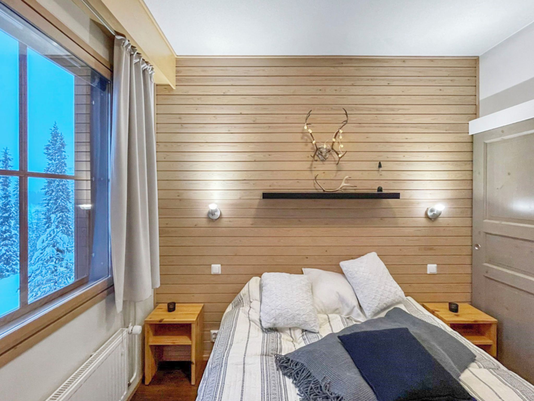 A bedroom with a wooden slat wall, a bed with pillows, a nightstand, a window with curtains, and a view of snow-covered trees.