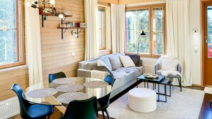 A cozy living room with a sofa, chairs, a glass table, and a coffee table. There are shelves with decorative items and a window with curtains.