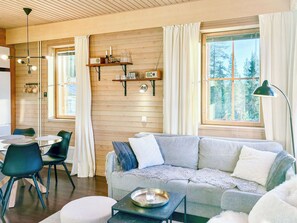 A cozy living room with a wooden wall, a sofa, a dining table with chairs, and a large window with curtains.