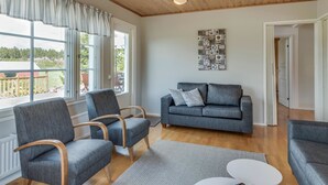 Ett vardagsrum med en grå soffa, två fåtöljer, ett träpanelat tak och ett stort fönster med utsikt över omgivningen.