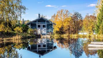 A house with a reflection in a calm body of water, surrounded by trees with autumn foliage.