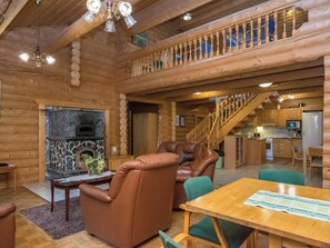 A log cabin interior with a stone fireplace, leather armchairs, a wooden dining table, and a kitchen area.