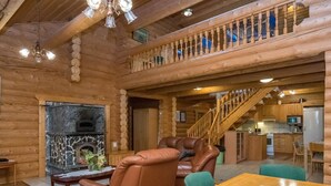 A log cabin interior with a fireplace, leather chairs, a dining table, and a kitchen area.