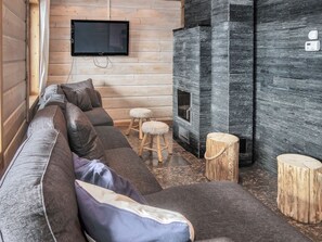 Interior de una cabaña de troncos con un sofá, dos taburetes, una chimenea y un televisor montado en la pared.
