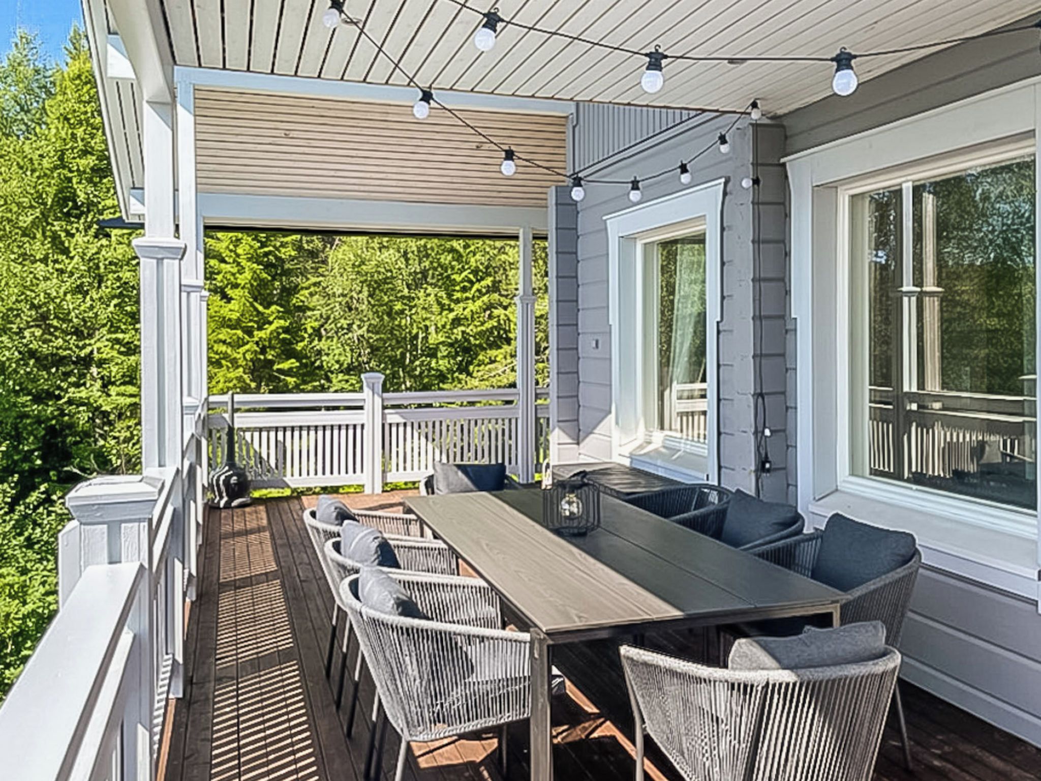 A covered outdoor deck with a dining table and chairs, surrounded by windows and a view of trees.