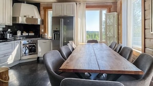 Een moderne keuken met witte kastjes, een grote houten eettafel en uitzicht op de omgeving door grote ramen.